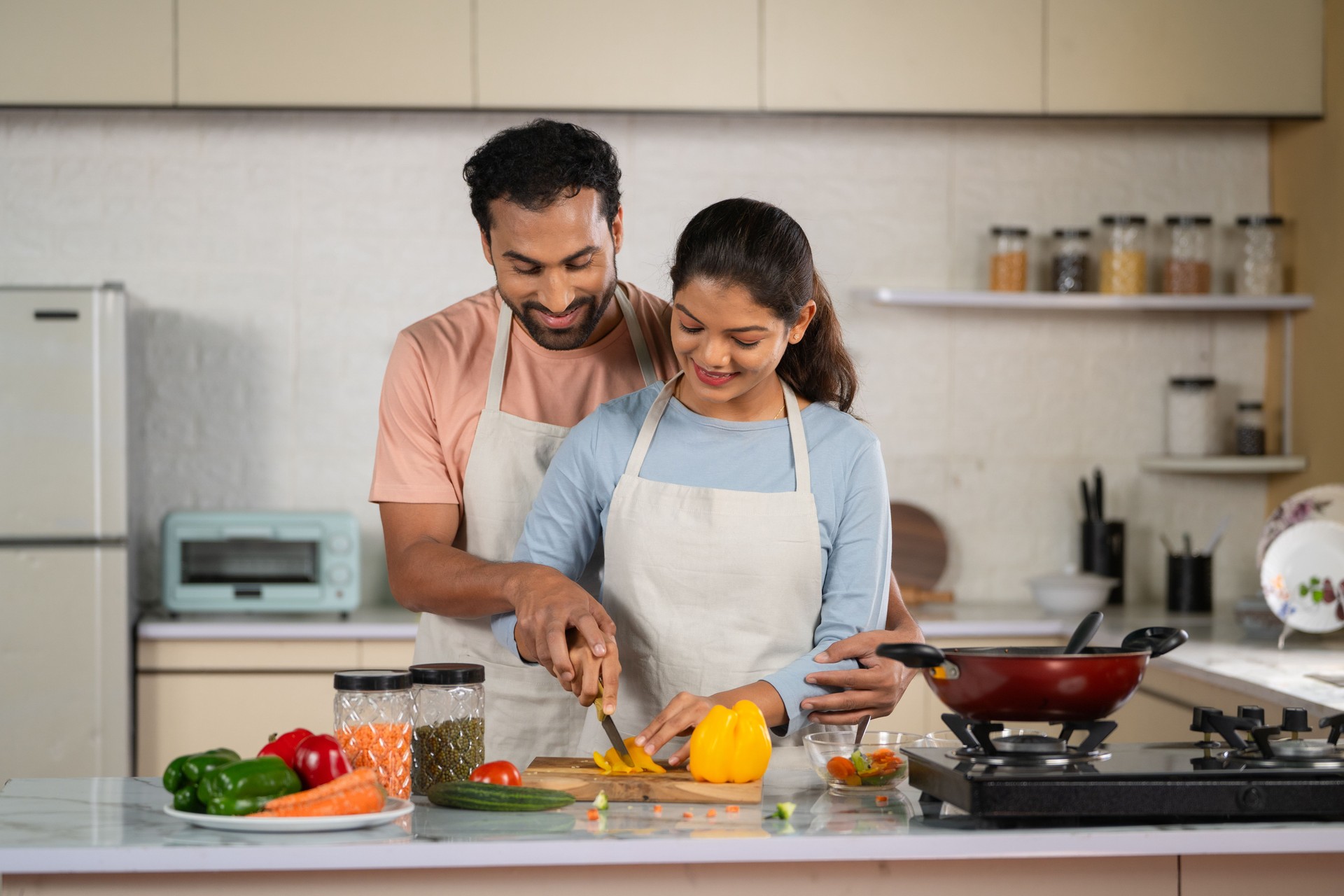 Romantic couples preparing food by slicing vegetables at kitchen while preparing food by hugging - concept of relationship affection, newlywed and togetherness